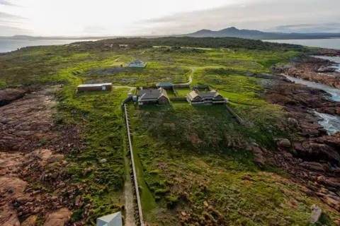 Nick Esser/Parks Victoria Aerial photo of small Gabo Island, with buildings visible, grassy, rocky shoreline, and see and land in the distance.