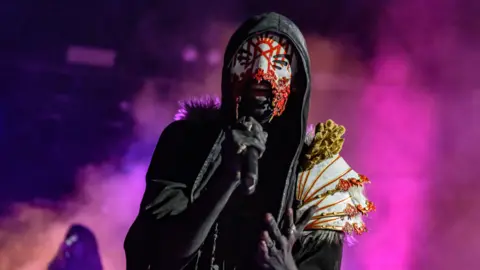 Getty Images A man singing into a microphone with a black hoodie on, wearing a red white and black mask - with white yellow and red shoulder pads.