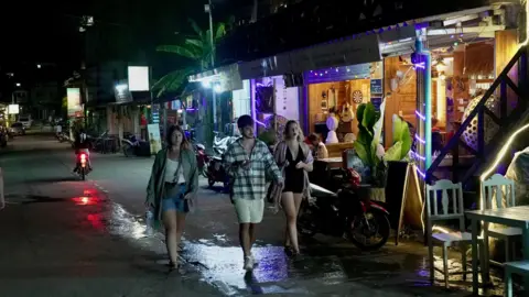 A group of three young people walk down a street in Vang Vieng with bars seen in the distance