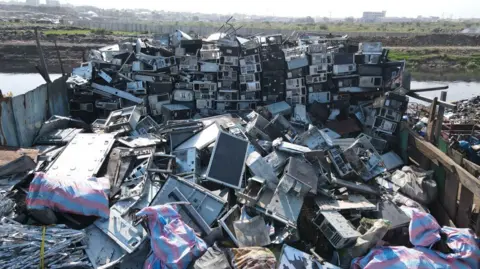 A huge pile of e-waste, including large screens, at the Agbobloshie dump. A body of water is visible in the background