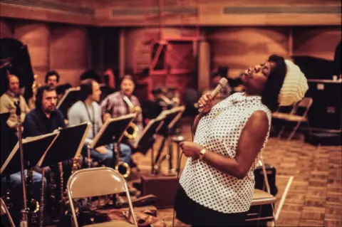 Getty Images Sarah Vaughn in full flight, performing with a live orchestra. She's wearing a cream woolly hat with a polka dot sleeveless blouse.