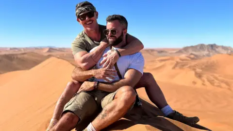 Luke Evans Luke Evans and his partner Fran Tomas cuddling on a sand dune. Luke Evans is behind wearing a khaki green t-shirt and matching headband along with black sunglasses. Fran Tomas is in front wearing a white T-shirt with khaki shorts and boots