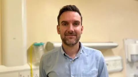 Sandwell and West Birmingham NHS Trust Dr Mike Blaber in a blue shirt in a hospital ward with equipment in the background