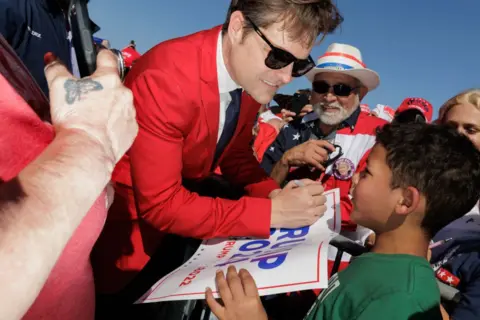 Getty Images US Republican Representative Matt Gaetz signs posters and hats at a Trump rally in Texas in March. He is smiling at a child holding a sign with 