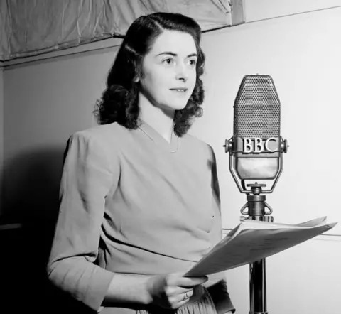 Black and white photo of SPencer standing in front of an old-fashioned BBC microphone holding a script