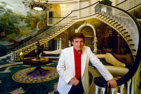 Getty Images Donald Trump, dressed in a red shirt and white blazer, stands in a huge, grand hallway in his mansion.