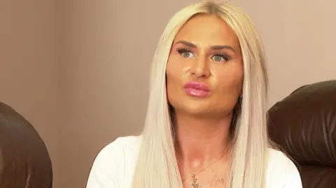 Samantha Morris sitting in her home looking thoughtful. She has long blonde hair and is wearing a white top. 