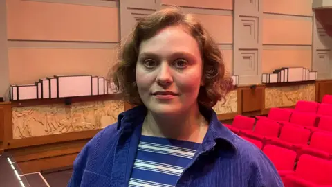 Thea Beyleveld is pictured in a radio theatre, with red seats behind her left shoulder while she wears a dark blue cord jacket and a blue and white striped T-shirt underneath