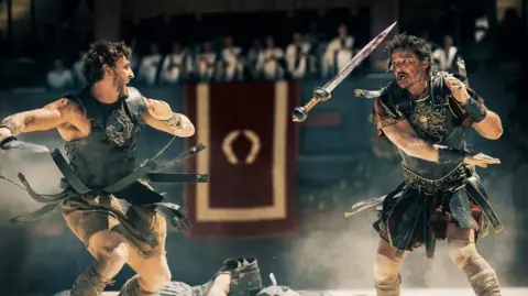 Paramount Pictures Two gladiators fighting in a still from the film Gladiator II, wearing traditional Roman clothing