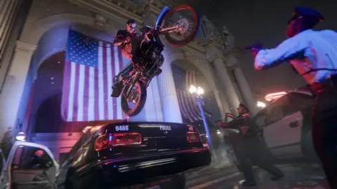 Activision-Blizzard Screenshot shows a man in leather jacket and shades on a motorcycle sailing through the air over the top of a parked police car. On the ground two police officers with pistols drawn raise their weapons, looking up at the bike as it flies above them. In the background a white-fronted building with two very large American flags hanging inside large arches is visible.