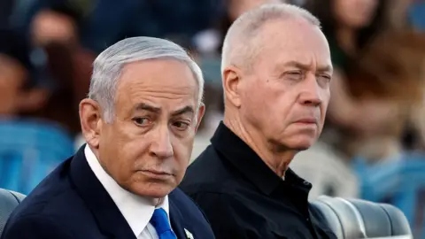 Reuters Benjamin Netanyahu, with combed over grey hair, wears a black suit, white shirt and blue tie and looks over his right shoulder as he sits next to Yaov Gallant who is wearing a black shirt and squinting