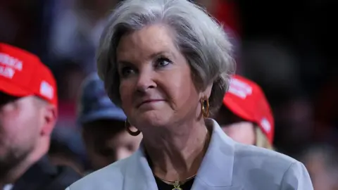 Reuters Susie Wiles, who has a grey cropped haircut, wears gold hoop earrings with a gold pendant necklace and a black top underneath a baby blue blazer which has a white and gold brooch on it as she looks on during a Trump rally