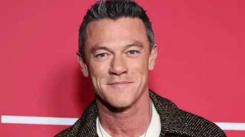 Getty Images Luke Evans smiling at the camera. He is standing in front of a red background with a white line just behind him. He is wearing a dark brown tweed jacket and a silver necklace. 