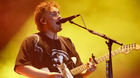 Getty Images Sam Fender performing on stage. He plays a black and white striped electric guitar while singing into a microphone. He wears a black T-shirt and has short curly fair hair. He's lit by a yellow spotlight.