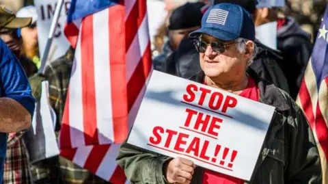 Getty Images A protester wearing a baseball cap with an American flag on it holds a placard reading 