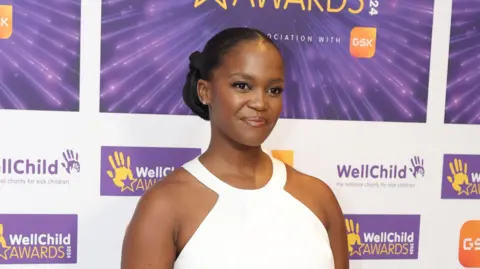 Chris Jackson/Getty Images Oti Mabuse wearing a white dress standing in front of backdrop promoting WellChild Awards