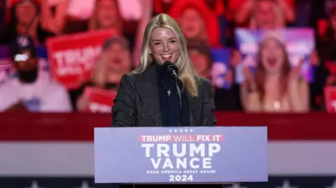 Reuters Pam Bondi addresses a crowd at a Trump rally. People are cheering and shouting behind her, holding Trump-Vance signs. Bondi has long blonde hair, and wears a grey blazer.