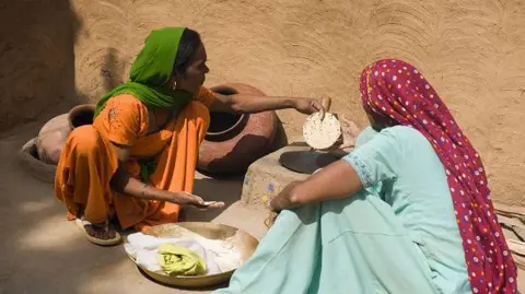 Getty Images Two women seen sitting in the sun as they make Indian flatbread on a stove. One of them is dressed in orange salwar-kameez with a green dupatta on her head. The other wears a pale blue salwar-kameez with a maroon dupatta with colourful dots.  