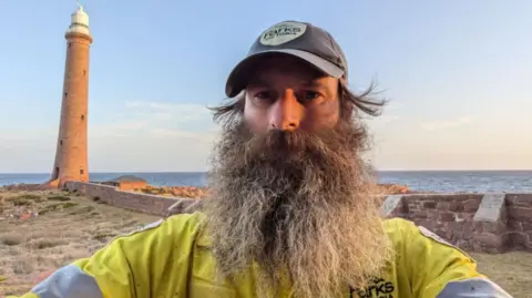 Sandy Duthie Sandy Duthie, man with large greying beard, yellow Parks Victoria jacket, with a lighthouse in the background, and sea, and blue skies.