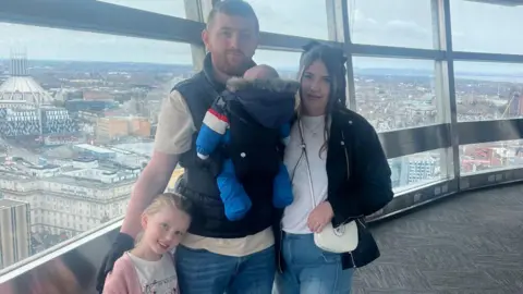 Ellie Bryan and Morgan Davies Morgan and Ellie smile for the camera on a viewing platform overlooking Liverpool. Morgan has a baby strapped to his chest and their daughter is standing beside him in a pink cardigan