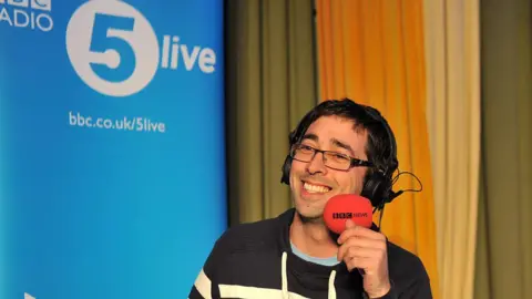 Colin Murray smiles as he presents on BBC Radio 5 Live. He is wearing black glasses and a navy and white jumper. 