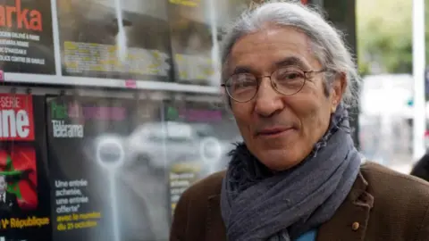 Getty Images Franco-Algerian writer Boualem Sansal poses in a brown jacket and grey scarf beside a newspaper kiosk in Paris in 2015