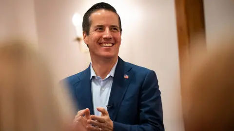 Getty Images Bernie Moreno claps and smiles as he looks away from camera wearing a blur suit with a US falg badge, in a white room