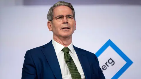 Getty Images Scott Bessent is seated during an interview and wearing a blue suit with a green tie and a white button-down shirt. He is wearing glasses.