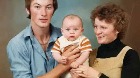 Luke Evans Luke Evans as a baby being held by his parents David and Yvonne. David is wearing a blue shirt, Yvonne is wearing a black polo-neck and a sandy coloured pair of dungarees. Luke in the middle is wearing a mustard and off-white babygro. 