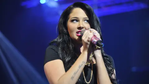 BBC/Sarah Jeynes Tulisa singing into a microphone. She is wearing a black t-shirt and has hot pink lipstick on.