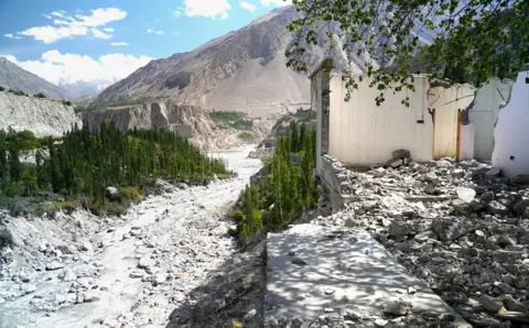 A damaged building with walls missing, and rubble on the floor, can be seen to the right of frame, with a river, trees and mountains to the left of frame. Picture shows the damaged village of Hassanabad.