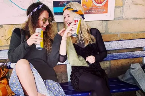 Rachel Adam Hazel laughing with a friend - they are both sitting on a blue bench and holding cans of lager 