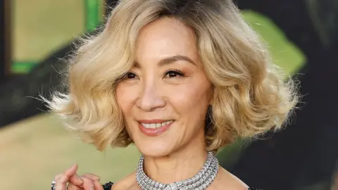 EPA Michelle Yeoh with blonde hair and a thick, sparkling necklace, in front of a green Wicked-themed background at the film's premiere in Los Angeles