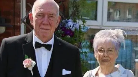 Family photograph Roy and Susan Pollit pose for a photograph together. Roy wears a formal black suit with a black bow-tie and a pink flower in his button hole. Susan  wears a white fascinator and a light grey outfit.