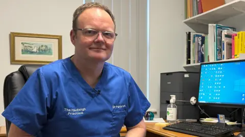 BBC A GP in his fifties wearing glasses in his consulting room. 