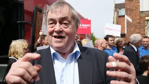 PA Media John Prescott out campaigning in Hull in 2017 with Labour Party supporters in the background. He is wearing a suit with a pale blue shirt and gesturing at the camera, he has a plaster on one finger. 