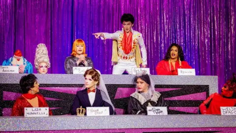 BBC/World of Wonder/Guy Levy Contestants compete in the Snatch Game. Sitting in two rows from L-R: Marmalade is a beheaded Marie Antoinette; Chanel O'Conor is Coco Peru; Kyran, dressed as Elvis Presley, stands and points at RuPaul; Rileasa is Rihanna; La Voix looks at Kyran while dressed as Liza Minnelli; Charra Tea is Nadine Coyle dressed as Mary Poppins; Lill is Queen Victoria and Actavia is the Welsh dragon. 