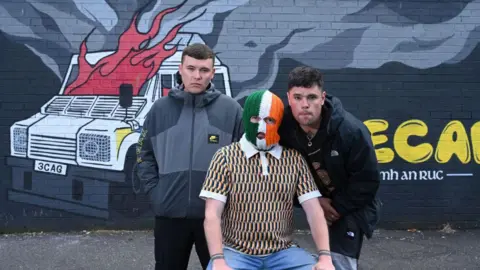 Michael Cooper Three men pose in front of a mural showing a burning PSNI vehicle. The man in the middle is wearing a tri-colour balaclava. The man on the left is wearing a grey jacket and the man on the right is wearing a black jacket.