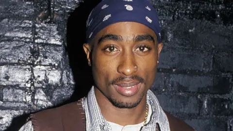 Getty Images Tupac Shakur standing against a black brick wall in 1994