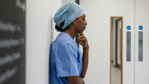 Getty Images An NHS hospital worker wearing scrubs and a hair net stands against a wall inside a hospital in Newcastle while looking pensive. Stock photo illustration.