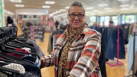 BBC/Simon Thake A woman in a colourful large checked coat and leopard-print scarf smiles for the camera in a charity shop.