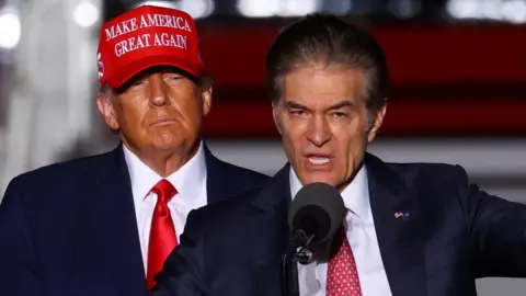 Reuters Mehmet Oz speaking into a microphone and gesturing as Donald Trump behind him looks on, wearing a red 