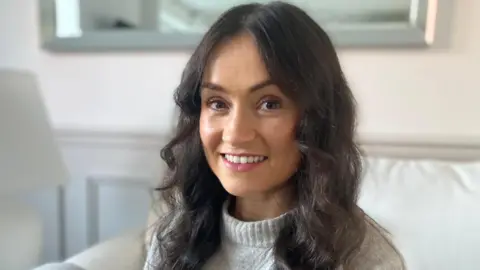 BBC A profile of Hannah Newby wearing a light grey woolly jumper and long, curly dark hair sitting on a sofa in front of a mirror.