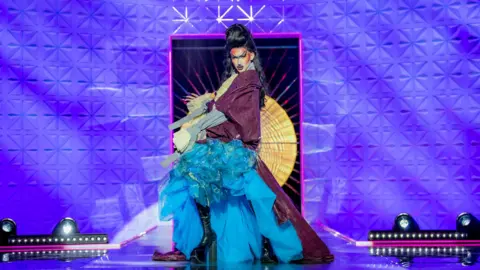 BBC/World of Wonder/Guy Levy Kyran Thrax walking the Drag Race UK runway in a purple and blue gown