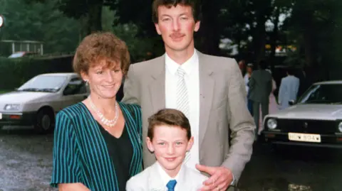 Luke Evans Luke Evans as a young boy with his parents Yvonne and David. Luke is in a pale grey suit and blue tie. David is in a grey suit, white shirt and striped tie on the right, and Yvonne is in a blue and black pinstriped jacket and black top, with pearls around her neck.