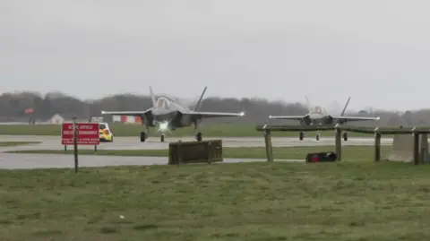 Shaun Whitmore/BBC Two planes, which appear to be F-35As from the Joint Strike Fighter programme, located at RAF Lakenheath. They are located on a runway or taxiway, surrounded by grass lawns. A police car is visible. 