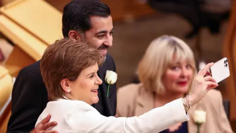 PA Media Nicola Sturgeon, with short brown hair and wearing a white suit, holds a white phone in front of her to take a selfie with Humza Yousaf, wearing a dark suit and standing next to Sturgeon with a hand on her back, in the Scottish Parliament. 