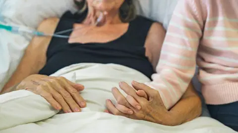 Getty Images A woman with tubes coming out of her nose lays on a bed and holds the hand of a child. Their faces are not visible.