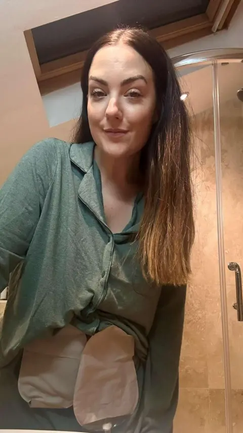 Anna Cooper Selfie of 31-year-old lady looking at the camera in a bathroom. She is wearing a green shirt and visible above it are two pale-coloured stoma bags. 