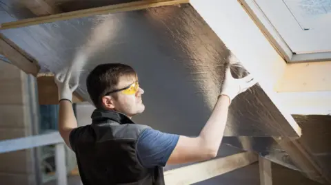 Getty Images A workman wearing safety specs fits some insulation between roof beams.